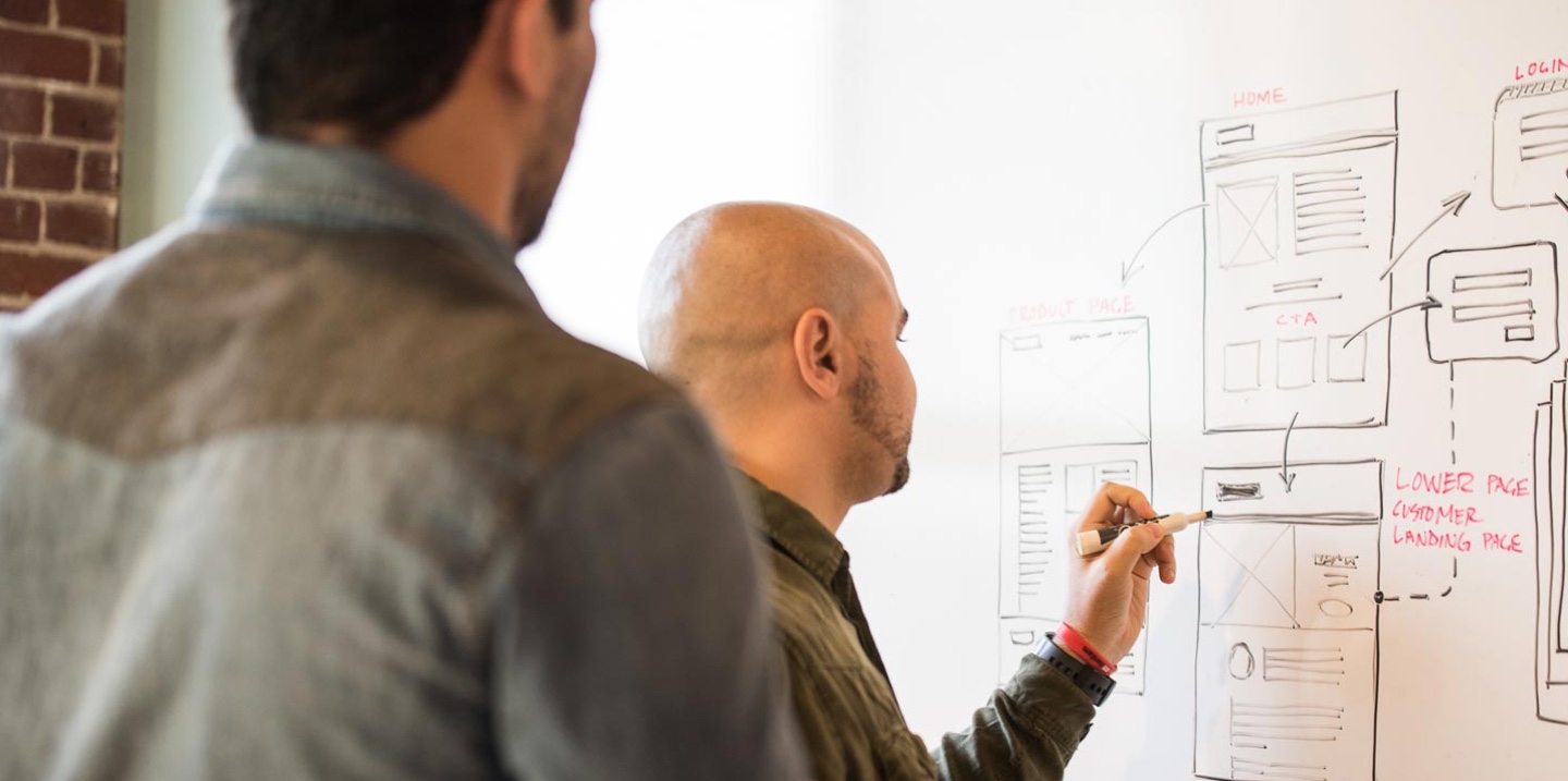 Photo of two web developers planning out a website in the first stages by sketching the necessary information and layout on a whiteboard. Concept Image - Decorative.